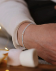 A delicate silver beaded bracelet with a central white pearl is shown on a person's wrist, wearing a cream-colored knit sweater. The background is softly blurred with warm fairy lights and neutral tones.