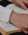 A delicate white beaded bracelet with a central pearl-like bead worn on a person's wrist, shown resting over an open book.