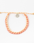 A braided cord bracelet in orange and cream, featuring a small silver-toned circular charm with a star design, displayed on a clean white background.