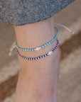 Two beaded ankle bracelets on a person's ankle, one in turquoise and white beads and the other in navy and white beads, with a gray denim cuff visible above.