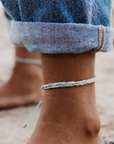 A close-up of a person's bare ankle wearing a thin braided rope anklet in pastel mint green, white, and light blue, with cuffed blue denim jeans visible above it. A second matching anklet is faintly visible on the other ankle in the background.