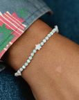 A close-up of a person's wrist wearing a beaded bracelet with alternating light blue and white beads and a small white star charm in the center. The person is wearing a denim sleeve with a colorful geometric pattern.