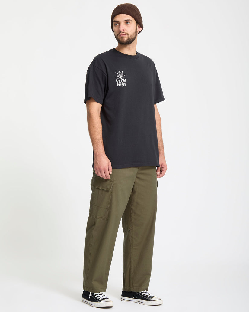 Man wearing a black Volcom t-shirt with a logo and green pants on a white background