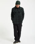 Person wearing a green plaid shirt, black pants, and a green beanie on a white background