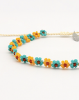 A beaded necklace featuring alternating yellow and turquoise flower beads with small orange centers, strung on a cream-colored cord with a silver heart-shaped clasp.