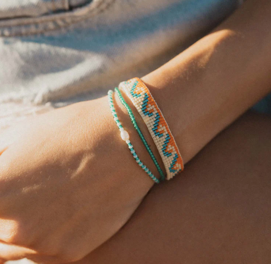 A close-up of a person's wrist wearing three bohemian-style woven bracelets. The bracelets feature geometric patterns in turquoise, orange, and cream, with one bracelet having a small white bead accent.