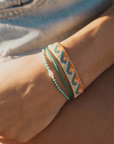 A close-up of a person's wrist wearing three bohemian-style woven bracelets. The bracelets feature geometric patterns in turquoise, orange, and cream, with one bracelet having a small white bead accent.