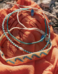 Three handmade beaded bracelets with turquoise, white, and orange details, displayed on a textured orange fabric outdoors.