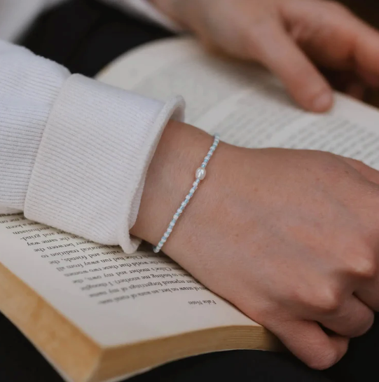 A delicate white beaded bracelet with a central pearl-like bead worn on a person's wrist, shown resting over an open book.