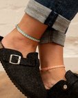 A close-up lifestyle shot of a person's feet wearing black felt slip-on clogs with silver buckles, paired with rolled-up blue jeans and two delicate braided anklets, set against a light brick wall and wooden floor.