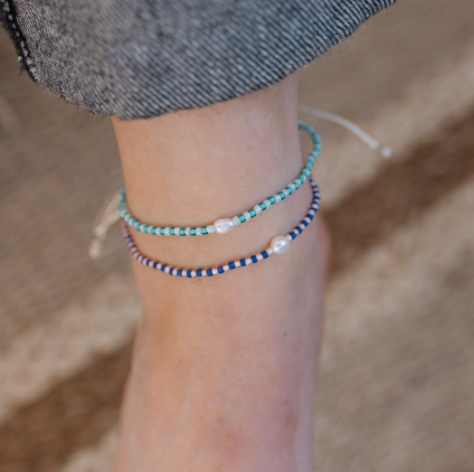 Two beaded ankle bracelets on a person's ankle, one in turquoise and white beads and the other in navy and white beads, with a gray denim cuff visible above.