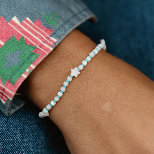A close-up of a person's wrist wearing a beaded bracelet with alternating light blue and white beads and a small white star charm in the center. The person is wearing a denim sleeve with a colorful geometric pattern.